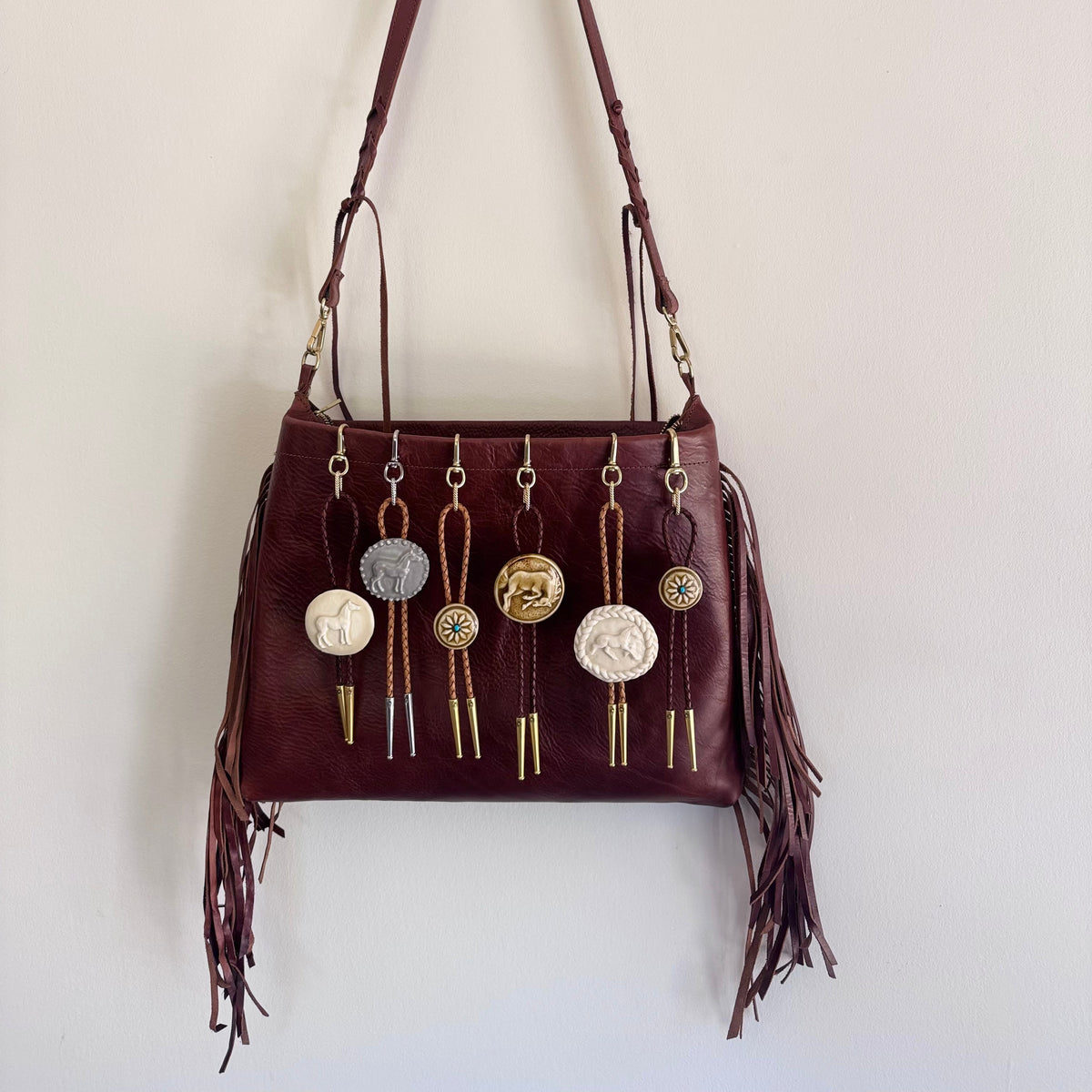 A dark burgundy leather shoulder bag with fringe details and multiple dangling bag bolo charms, hanging against a plain off-white wall.